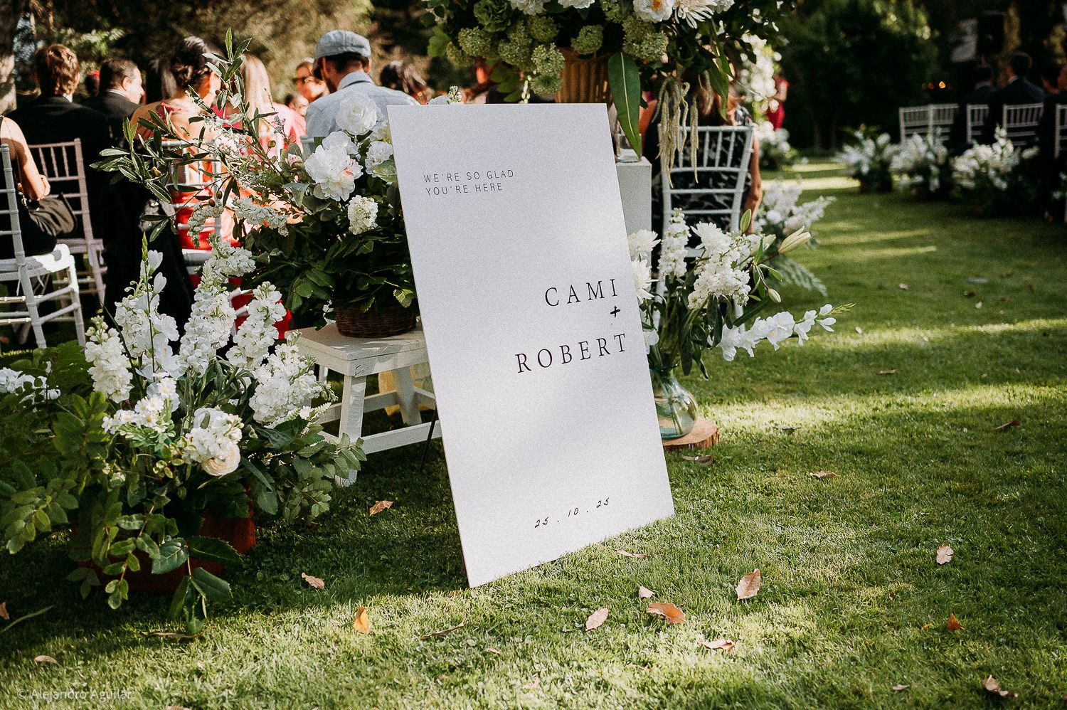 Elegant outdoor wedding ceremony sign for Cami and Robert, surrounded by lush floral arrangements on a green lawn.