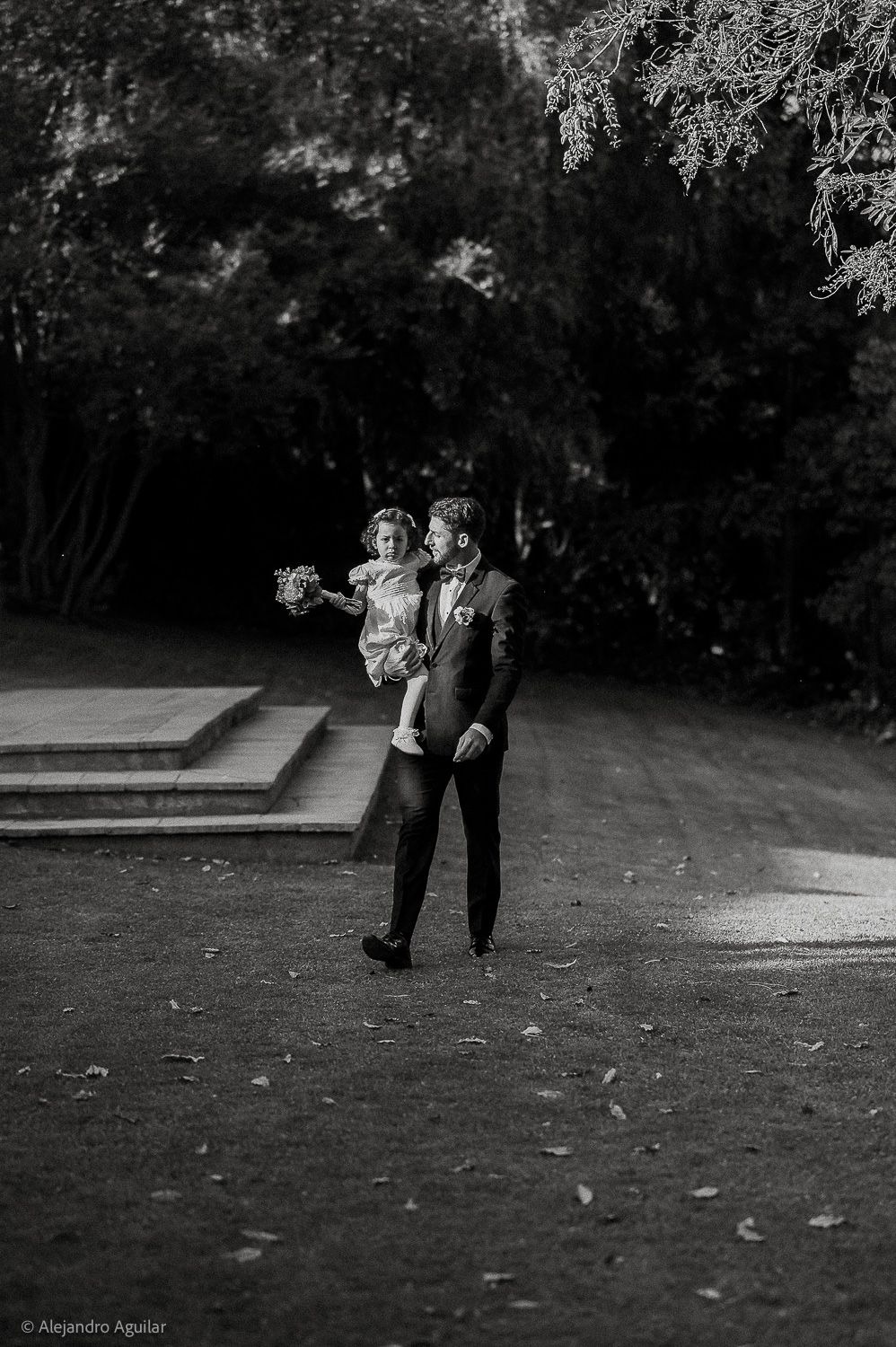 Black and white wedding photo of groom lifting bride outdoors, surrounded by lush trees, captured by Alejandro Aguilar.