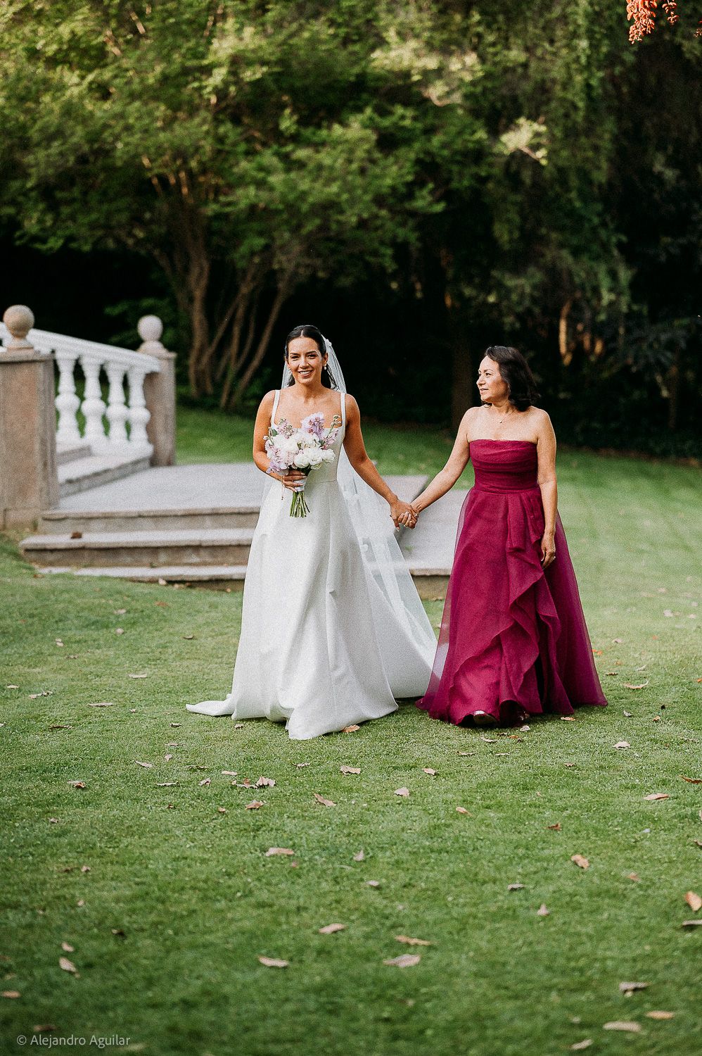 Bride in white gown holds hands with bridesmaid in burgundy dress on green lawn at outdoor wedding venue.