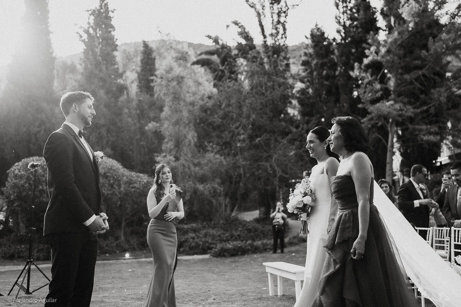 Black and white outdoor wedding ceremony with groom facing bride and bridesmaid, lush trees in background.
