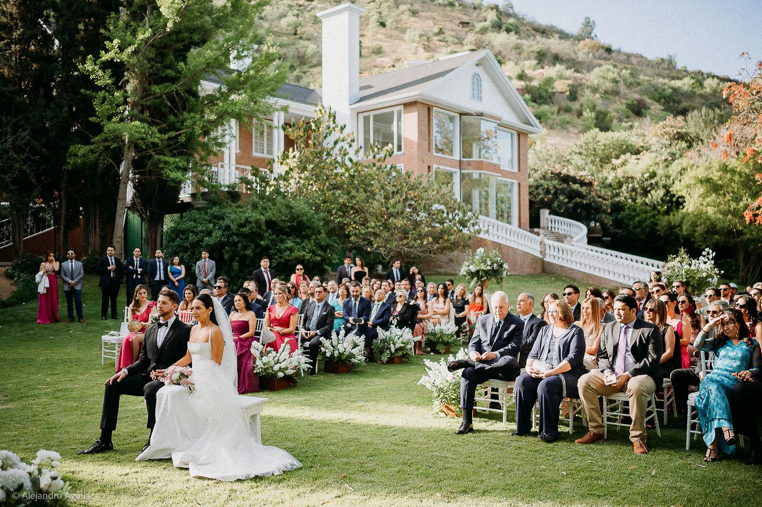 Outdoor wedding ceremony on lush green lawn with bride and groom seated, guests in chairs, and elegant brick mansion backdrop.