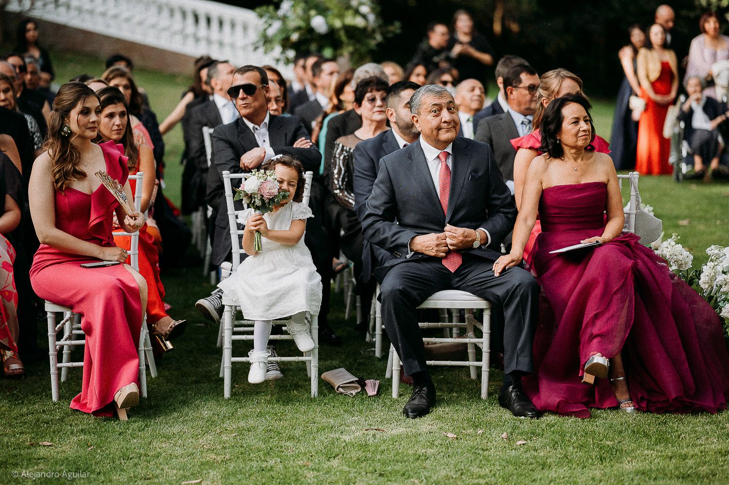 Guests seated outdoors at a wedding ceremony, including a flower girl in white dress and attendees in red and burgundy gowns.