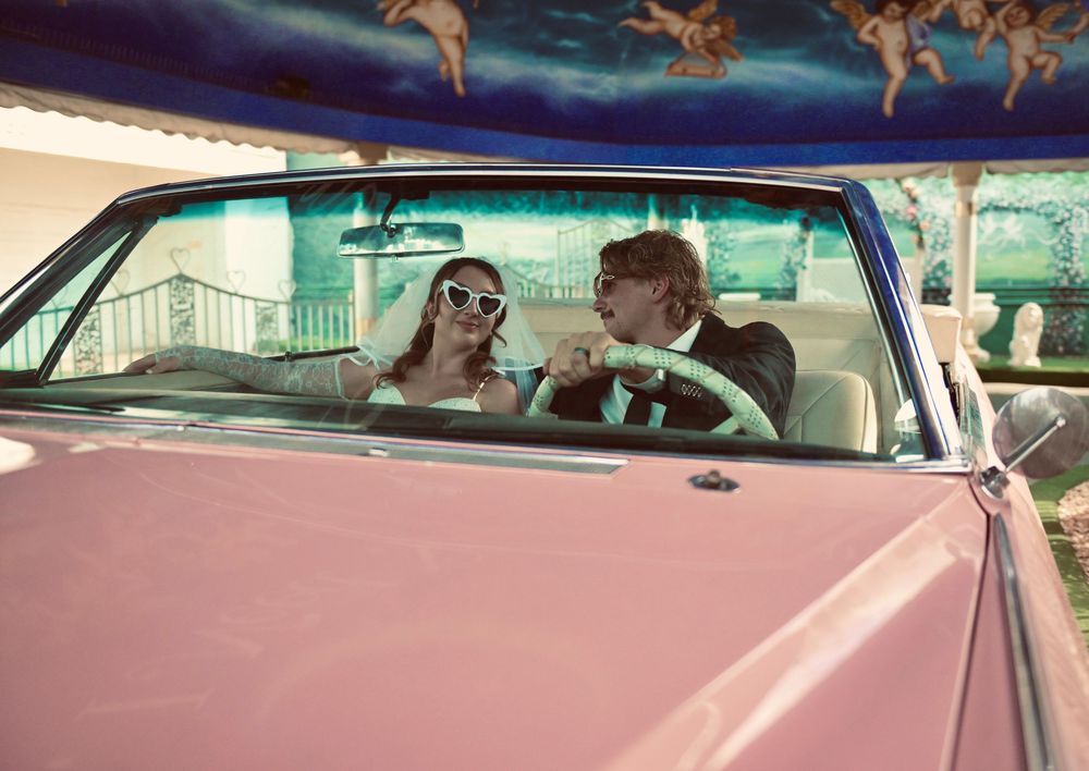 Newlywed couple in a pink convertible car, bride wearing sunglasses, groom in suit, Vegas wedding backdrop.