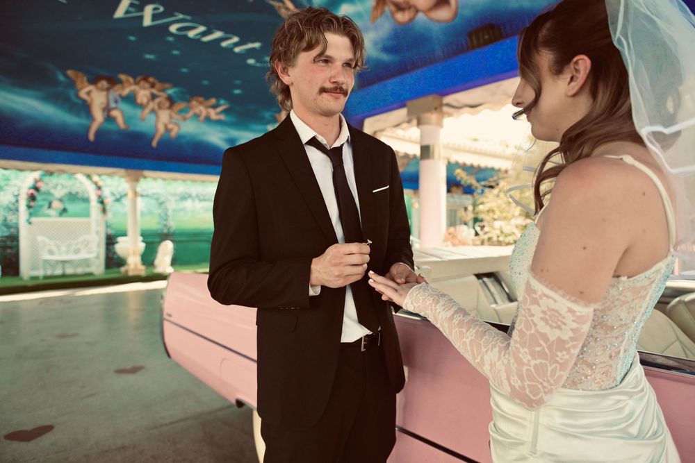 Couple in wedding attire exchanging rings at a colorful venue, groom in black suit with mustache, bride in lace dress.