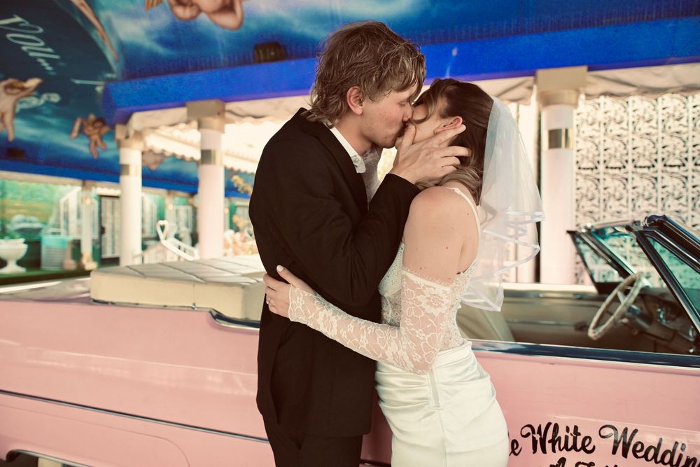 Newlywed couple sharing a kiss at a Las Vegas White Wedding chapel with a pink classic car backdrop.