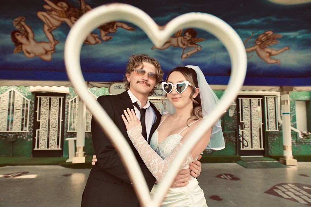 Newlywed couple posing through a heart-shaped frame, bride in white gown with heart sunglasses, groom in black tuxedo.