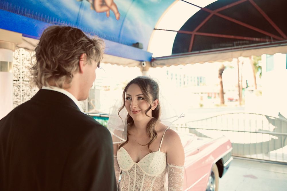 A bride in a white corset gown smiles at a groom in a black suit near a colorful carousel backdrop.