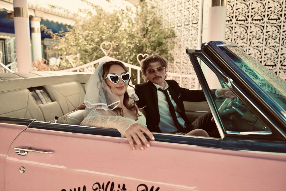 Newlywed couple in a pink convertible car, bride wearing heart sunglasses and veil, groom in black suit.