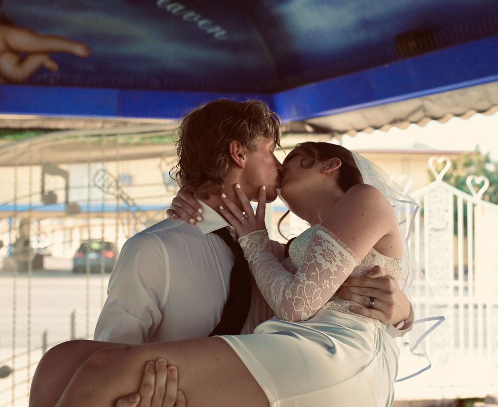 A groom carries his tattooed bride in a white dress as they share a romantic kiss outdoors.
