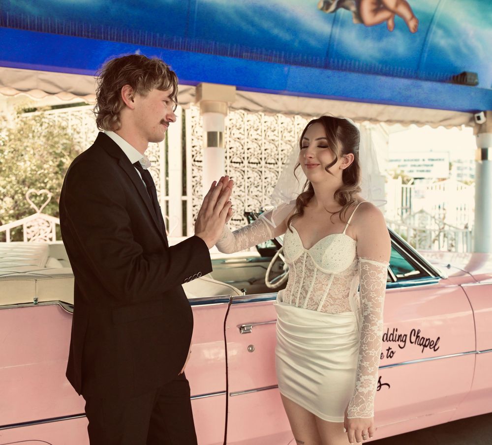 Couple in wedding attire poses by a pink car at the Little White Wedding Chapel in Las Vegas.