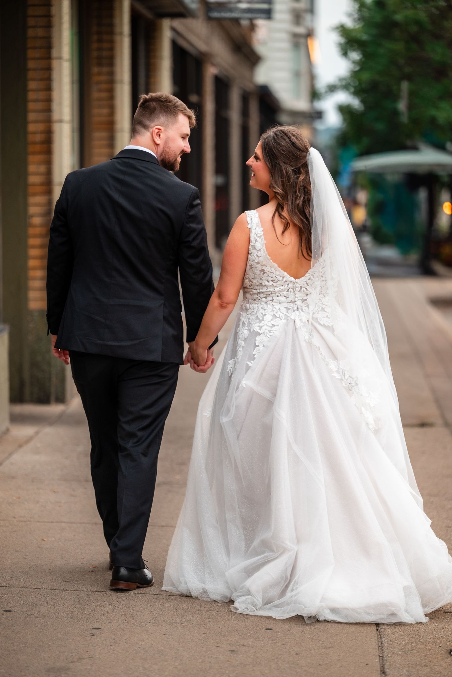Bride and groom walking hand in hand on a city sidewalk, bride in lace gown with veil, groom in black suit.
