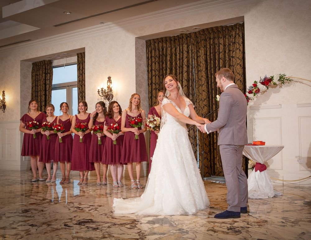 Bride and groom hold hands at indoor wedding ceremony with bridesmaids in burgundy dresses standing behind them.