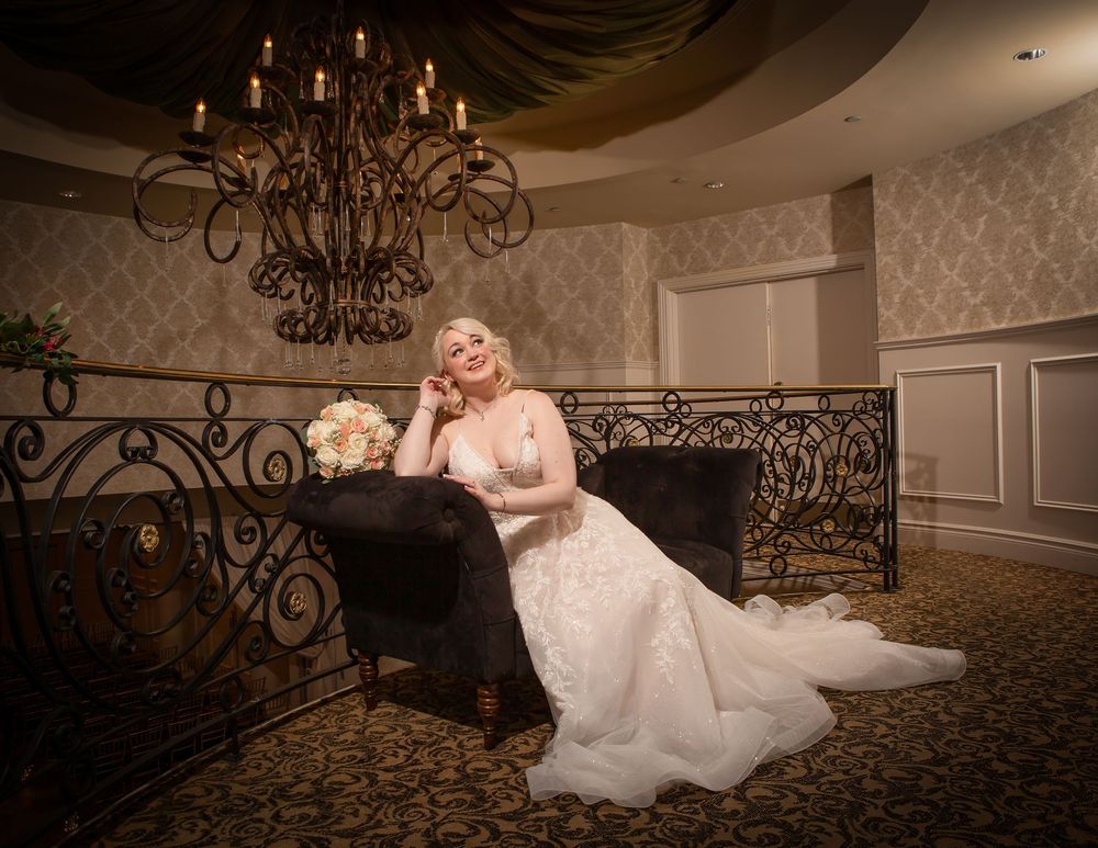 Elegant bride in lace wedding gown seated on ornate sofa under chandelier in luxurious ballroom venue.