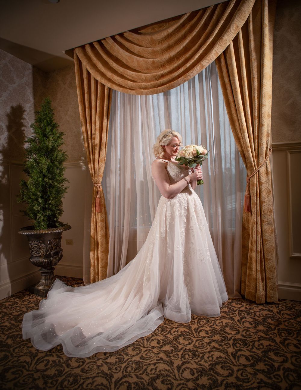 Bride in elegant lace ball gown holding bouquet stands before golden drapes in luxurious wedding venue.