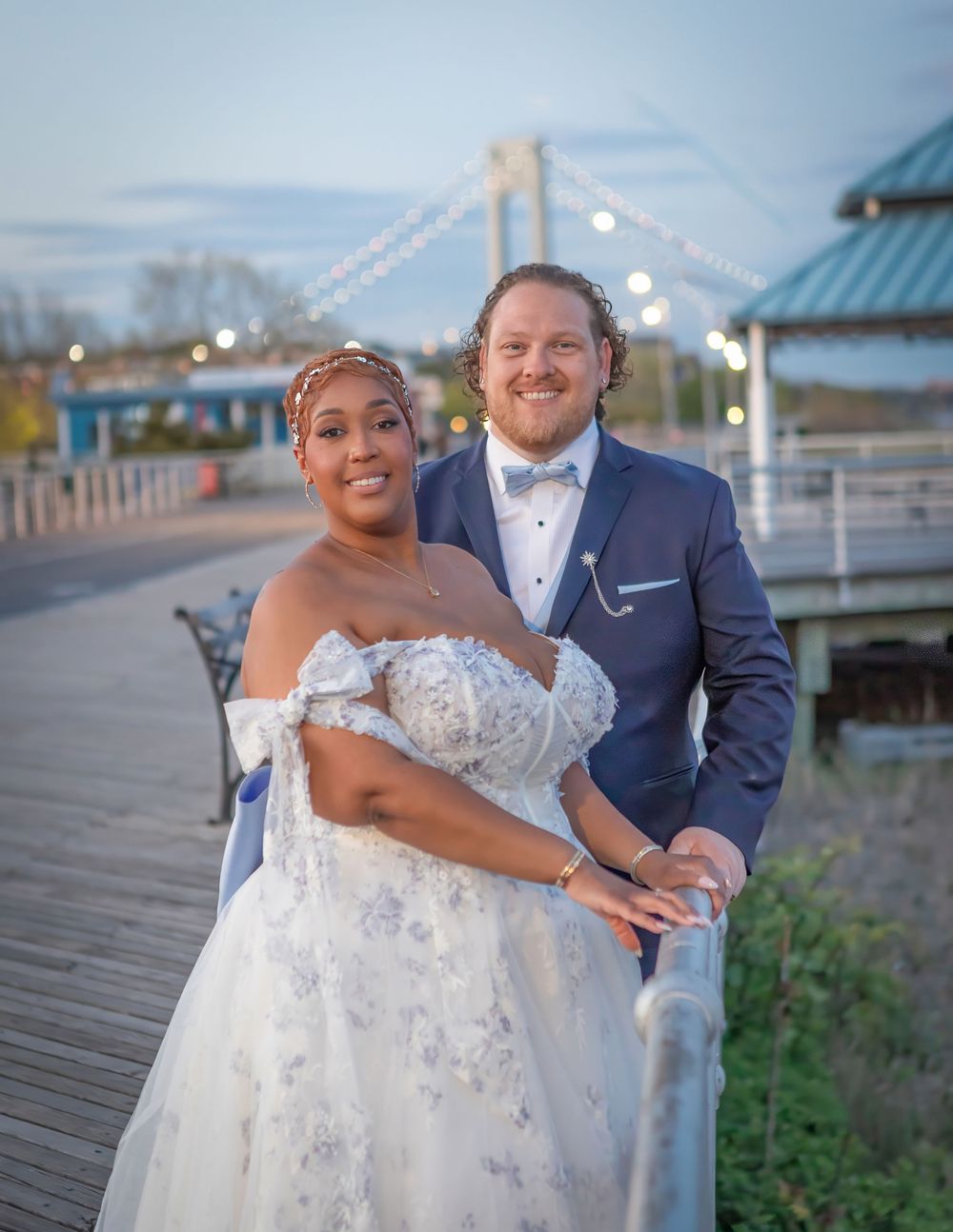 A couple in wedding attire poses on a waterfront boardwalk at dusk, bride in white gown and groom in navy suit.