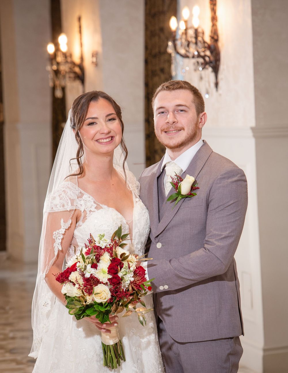 A smiling bride in lace gown holding red and white bouquet stands beside groom in gray suit at elegant wedding venue.