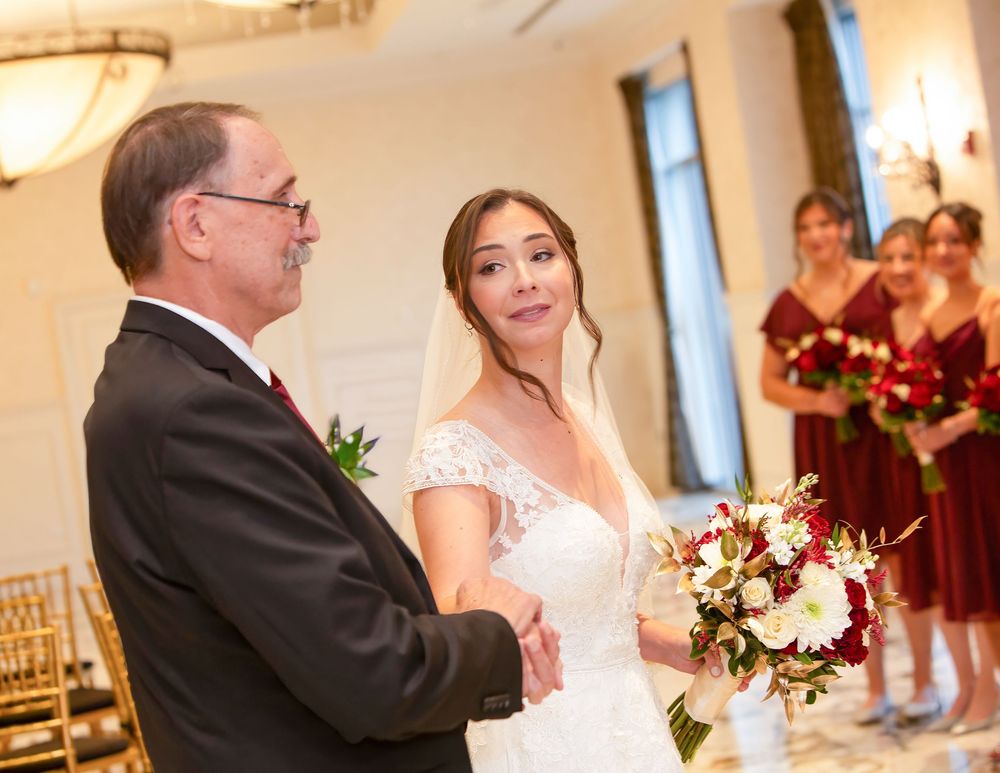 Bride in white lace gown holds red and white bouquet while sharing a moment with older man at elegant wedding ceremony.