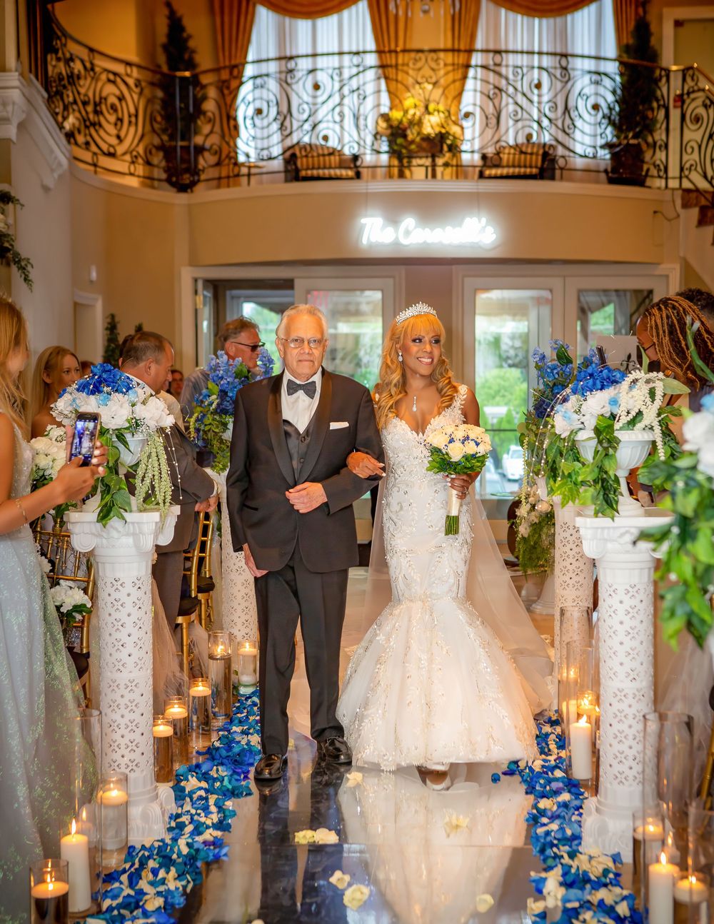 Bride in mermaid gown walks down candlelit aisle with escort, flanked by blue floral arrangements at elegant wedding venue.
