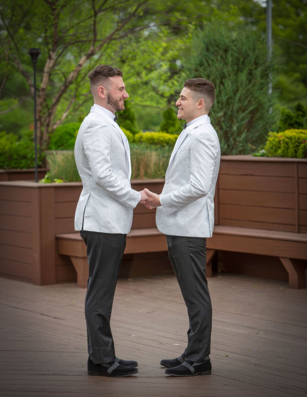 Two grooms in white blazers hold hands and face each other outdoors on a wooden deck surrounded by greenery.