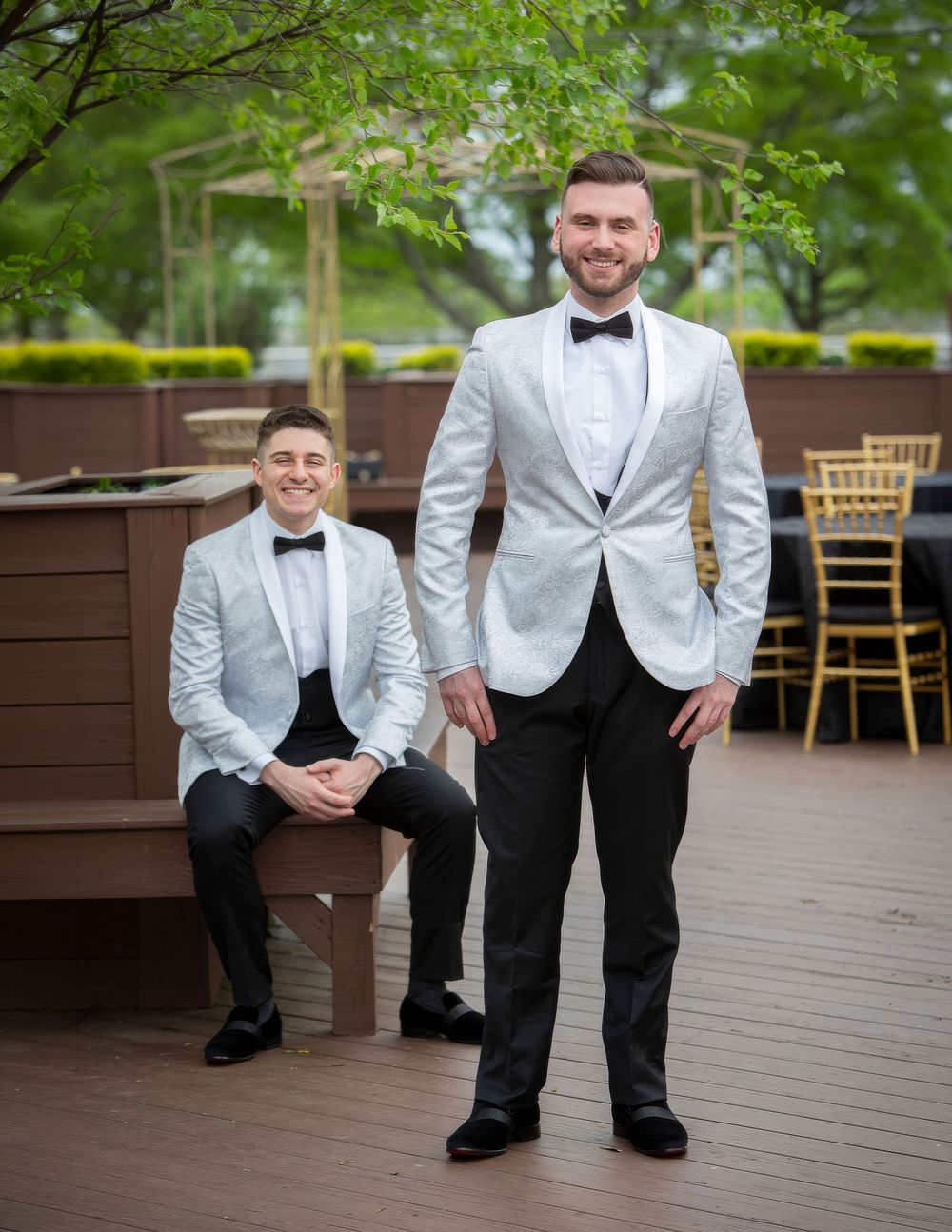 Two grooms in matching silver tuxedo jackets and black bow ties pose on an outdoor deck at their wedding reception.