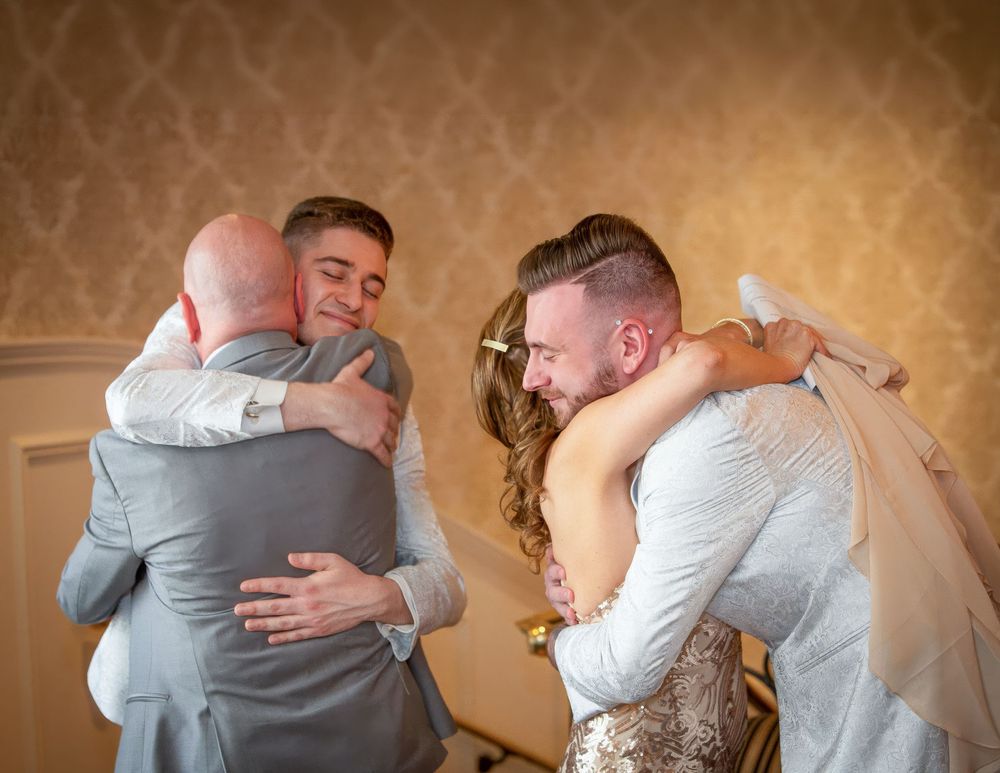 Bride and groom share emotional hugs with family members in elegant gold-wallpapered venue during wedding celebration.