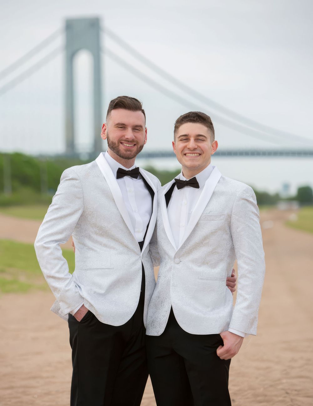 Two grooms in white tuxedo jackets and bow ties pose smiling near a suspension bridge on their wedding day.