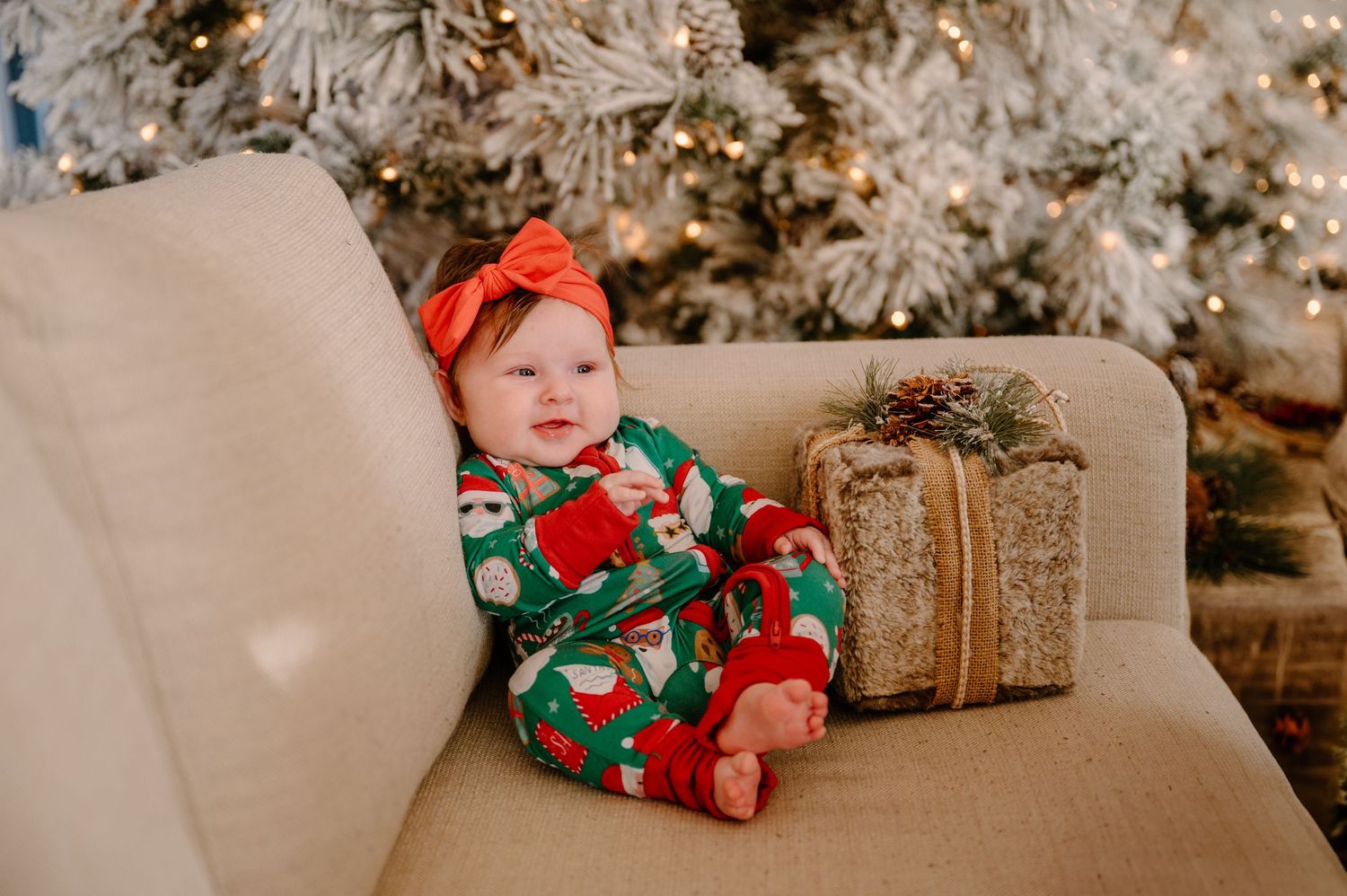 Baby in Christmas pajamas and red bow sits on a chair beside a gift box near a decorated holiday tree.