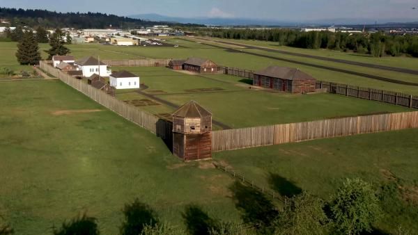 Aerial view of a historic wooden fort with watchtower, surrounded by green fields and a wooden fence in the Pacific Northwest.
