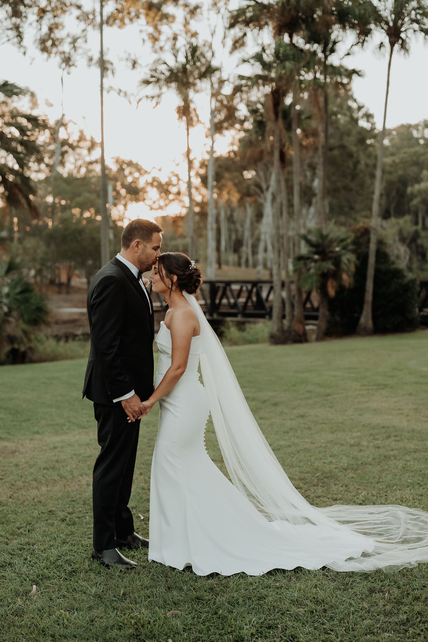 Bride and groom share a kiss on lush green lawn surrounded by tall palm trees during golden hour wedding portraits.