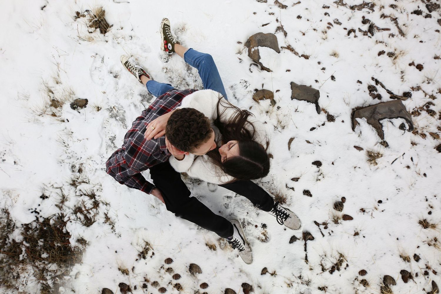 An overhead view of a couple lying close together in the snow, dressed in plaid and jeans, with patches of dirt showing.