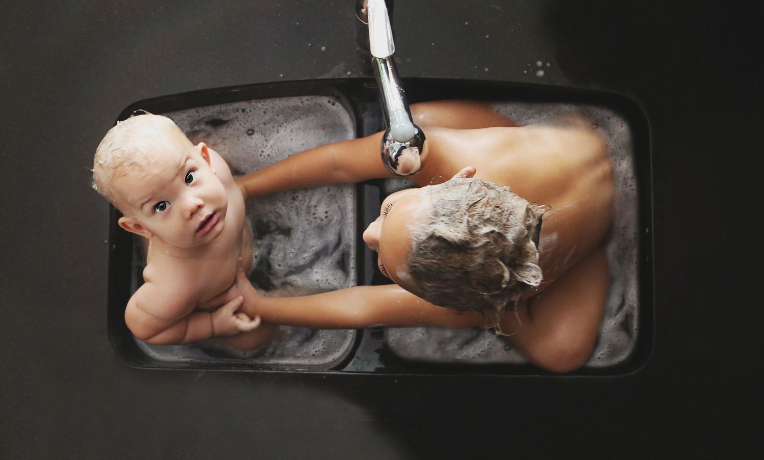 Overhead view of a mother bathing a smiling baby in a kitchen sink as water runs from the faucet.