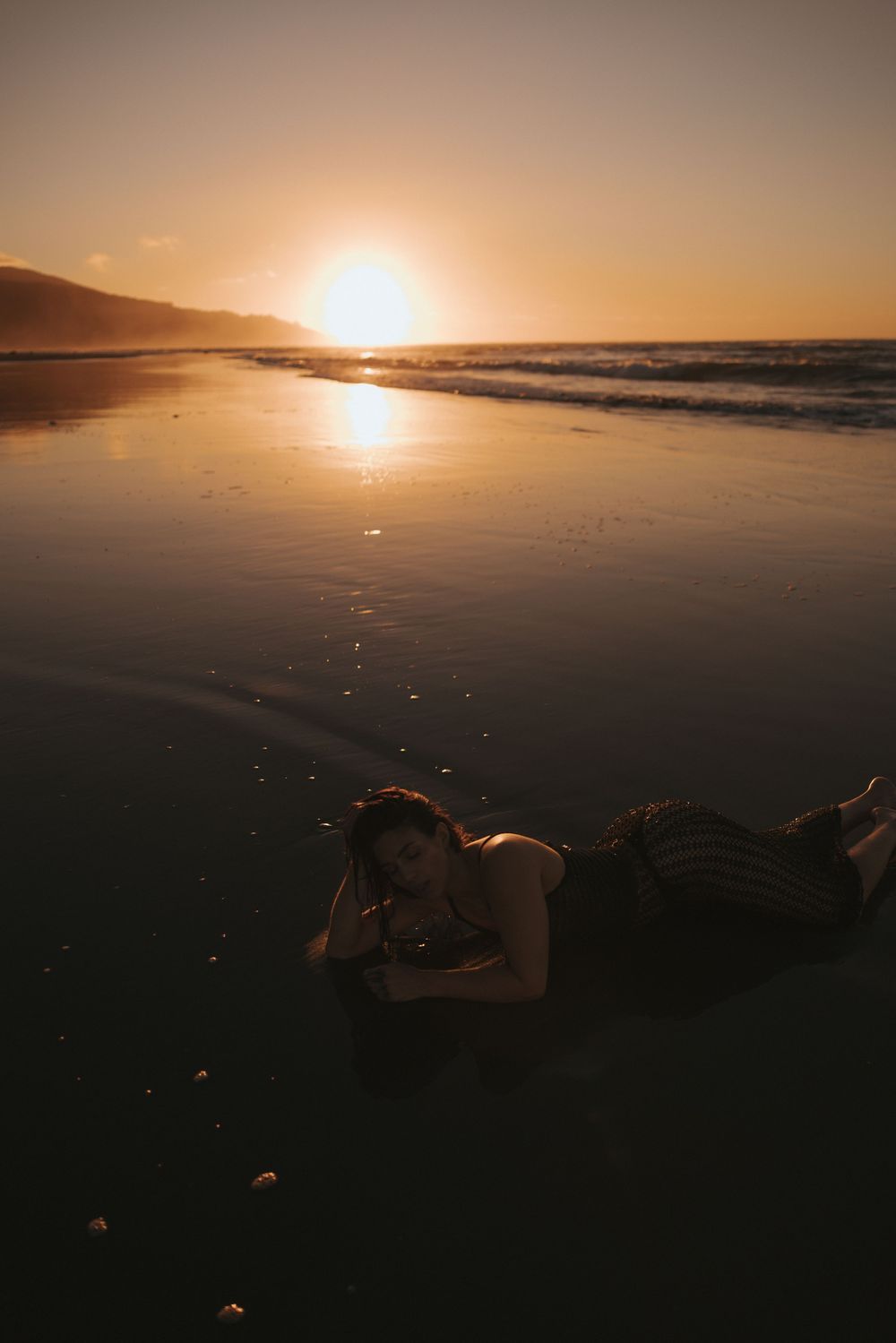 Woman in striped dress lying on reflective wet sand during golden sunset at beach in New Zealand..