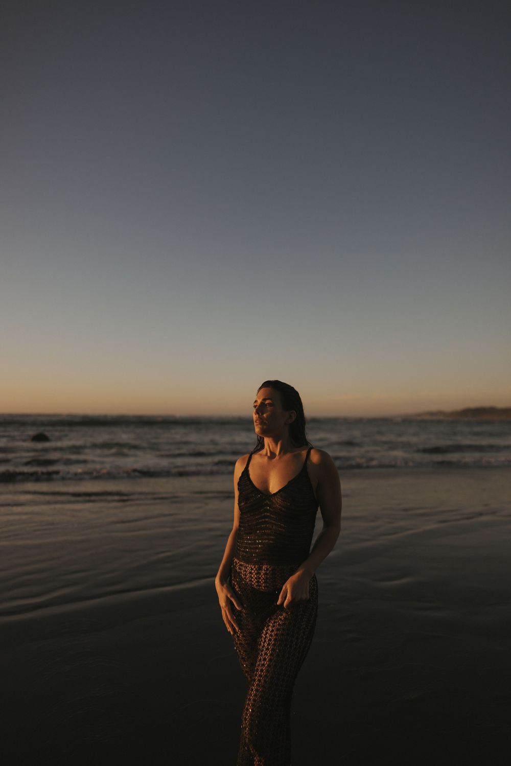 Woman in crochet dress standing on wet sand beach during twilight with eyes closed in New Zealand..