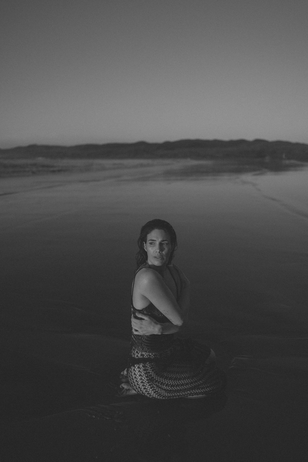 Black and white photo of woman in crochet skirt sitting on reflective beach at dusk in New Zealand..