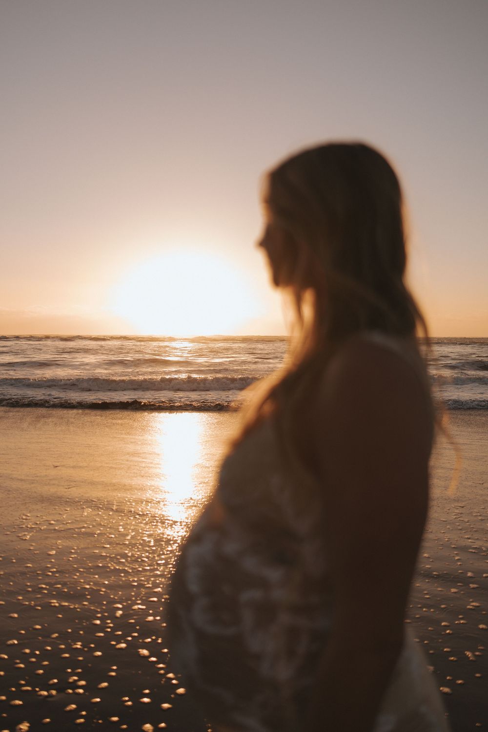 Silhouette of pregnant woman in floral dress gazing at golden beach sunset in New Zealand..