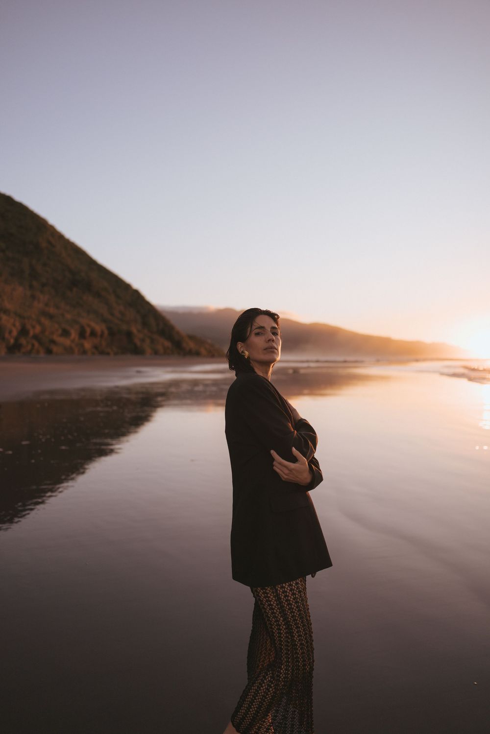 Woman in black blazer and patterned pants poses on reflective beach at golden sunset in New Zealand.