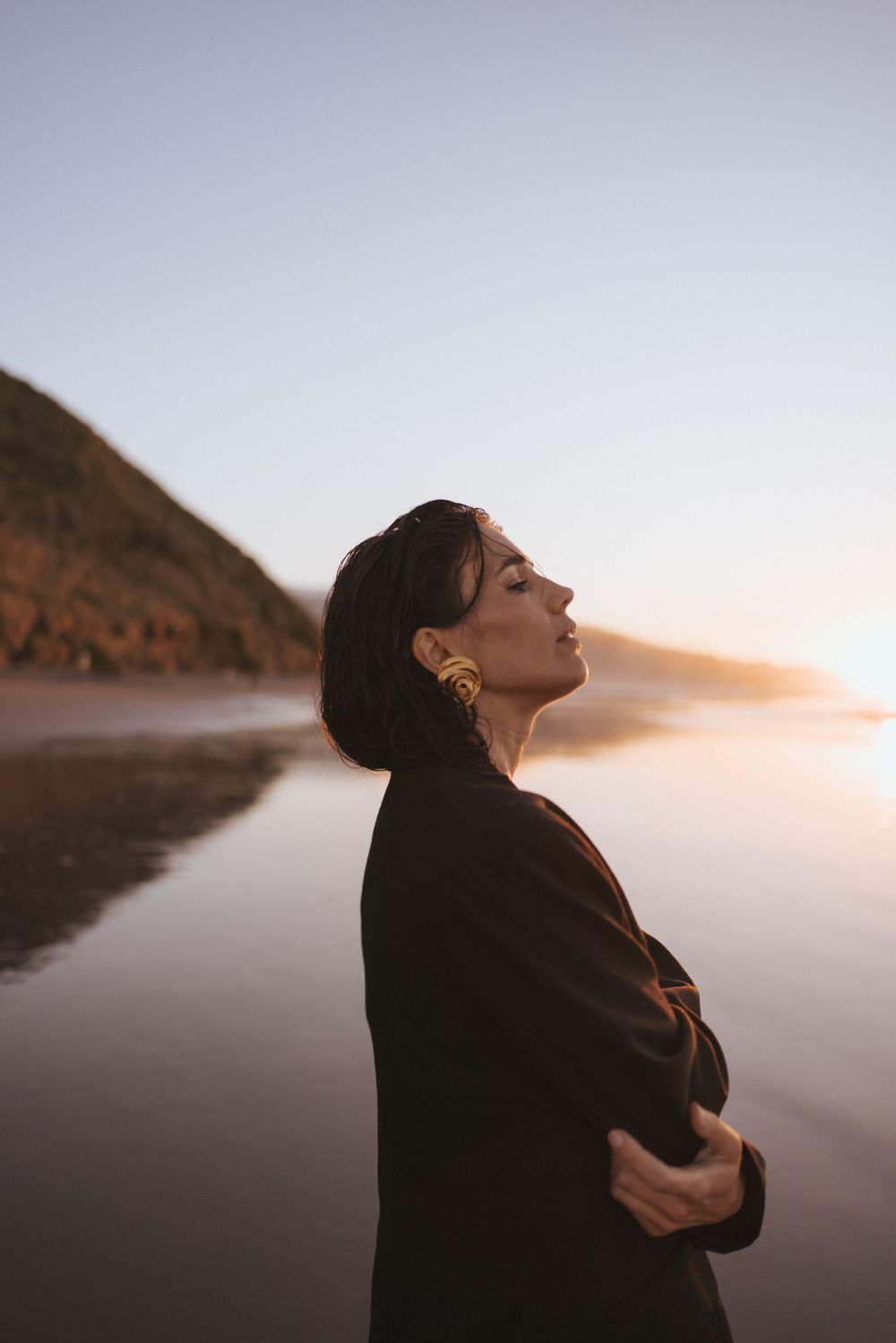 Woman in black blazer with gold earrings stands on reflective New Zealand beach at warm sunset.