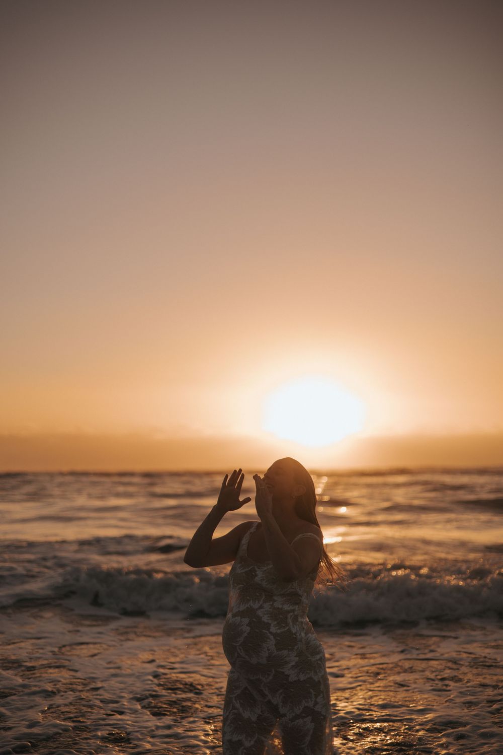 Pregnant woman in floral dress facing ocean waves during vibrant golden sunset in New Zealand..
