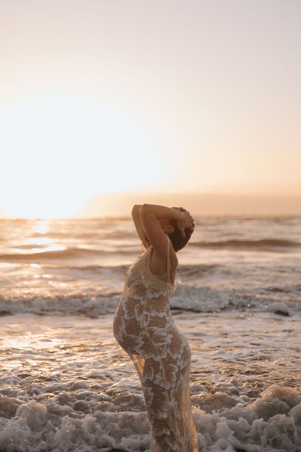 Pregnant woman in lace floral gown standing in ocean surf at sunset in New Zealand..