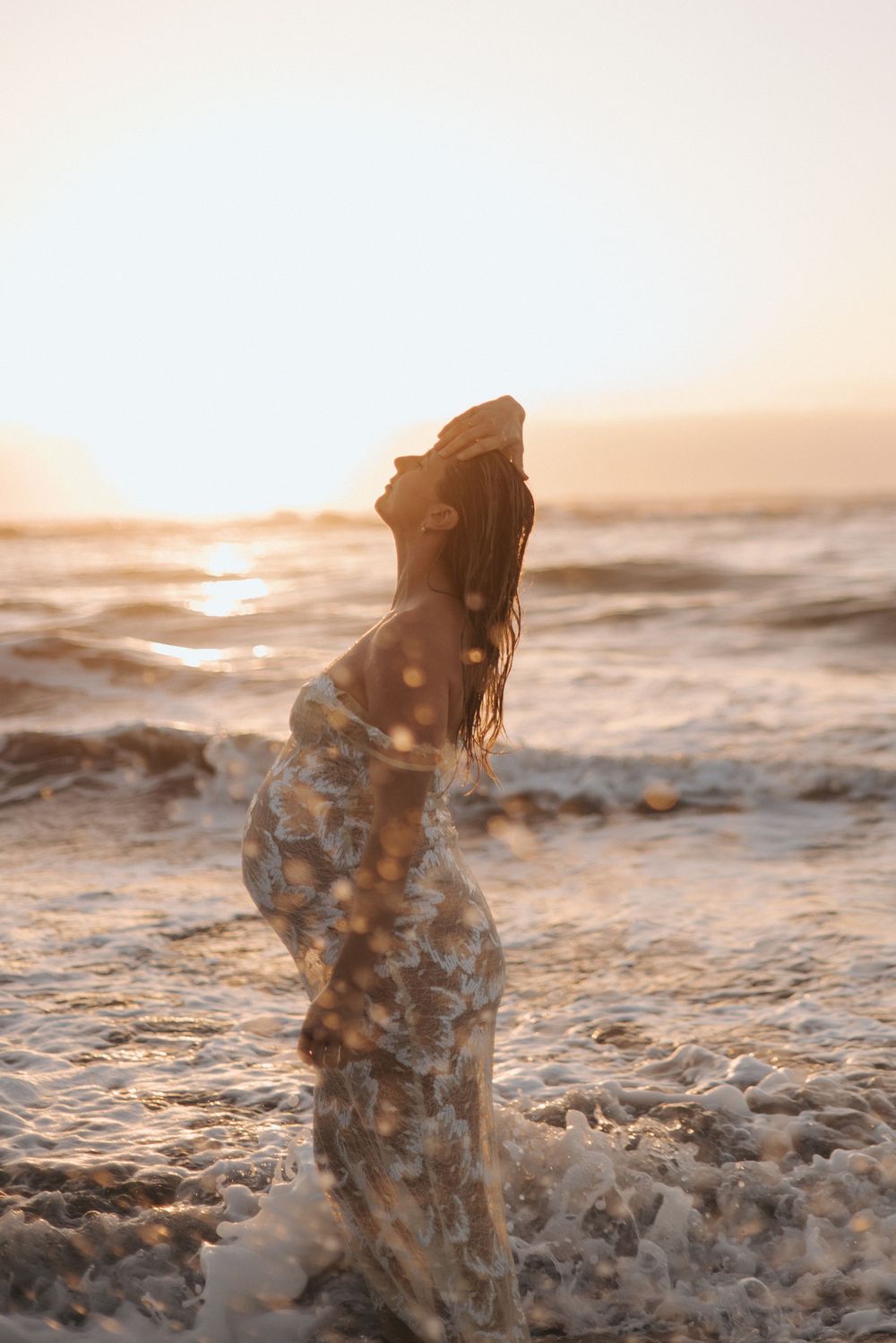 Pregnant woman in floral dress standing in sparkling ocean waves at sunset in New Zealand..