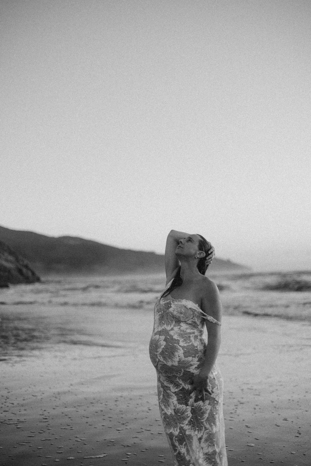 Black and white portrait of pregnant woman in floral dress on beach at dusk in New Zealand..