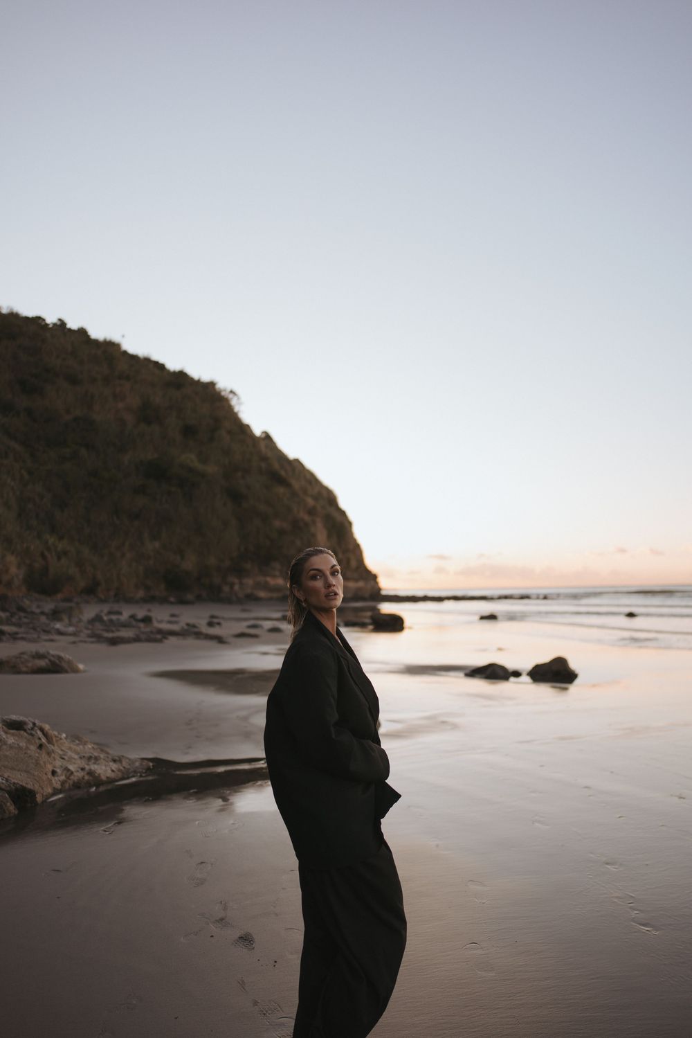 Woman in black suit stands on sandy beach at sunset with rocky cliff backdrop in New Zealand.