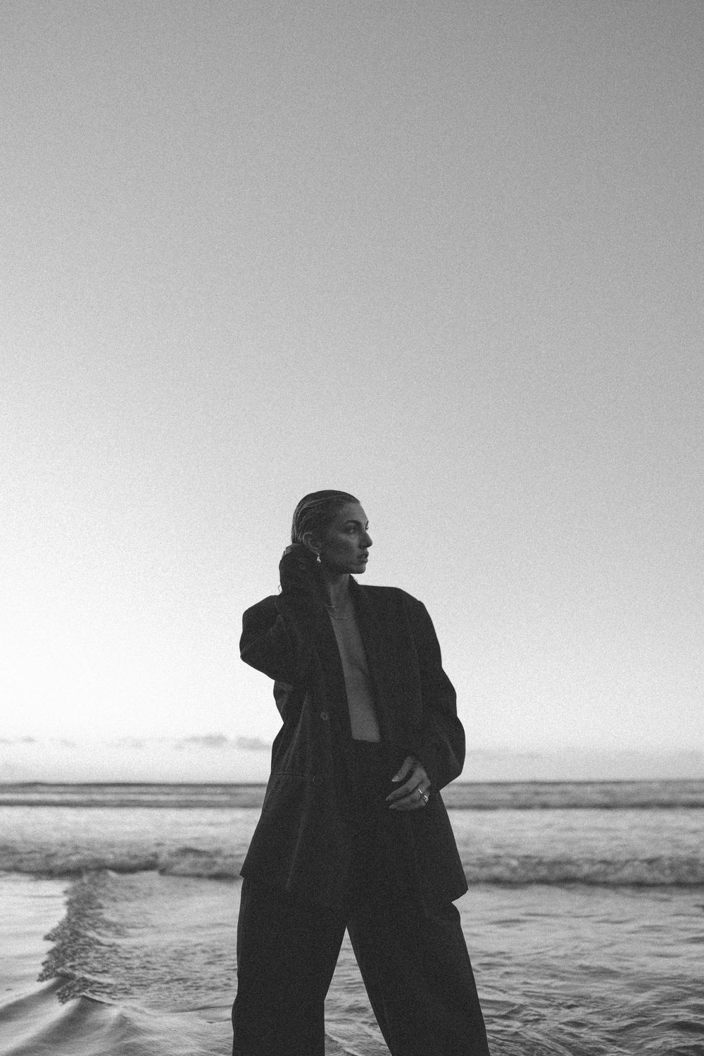 Black and white photo of woman in oversized blazer standing in ocean waves at sunset in New Zealand..