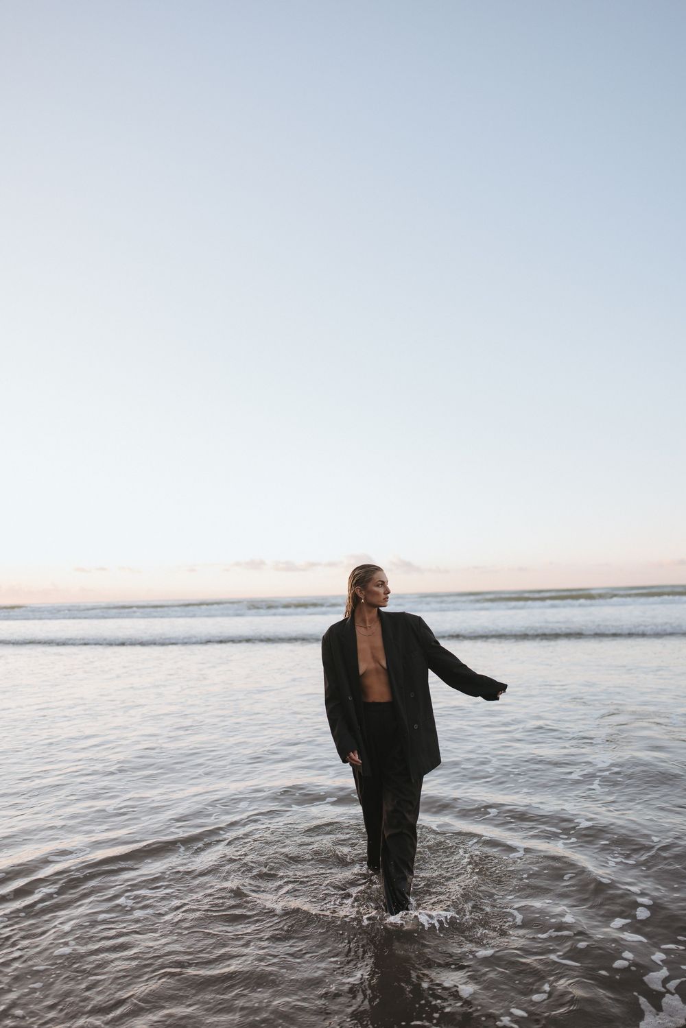 Woman in black blazer wading through shallow ocean waves on a serene beach at dusk in New Zealand..