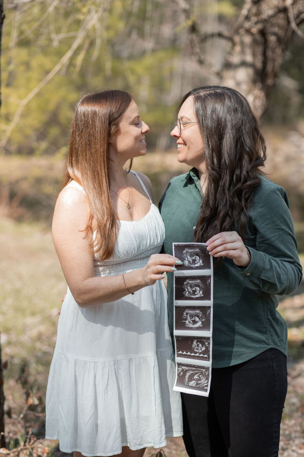 Two women share a joyful moment outdoors holding ultrasound photos, celebrating a pregnancy announcement together.