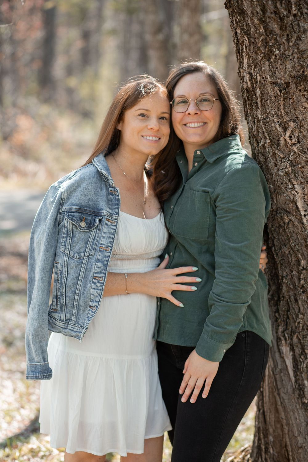 Two smiling women pose together near a tree in a forest, one in a white dress with denim jacket, other in green shirt.