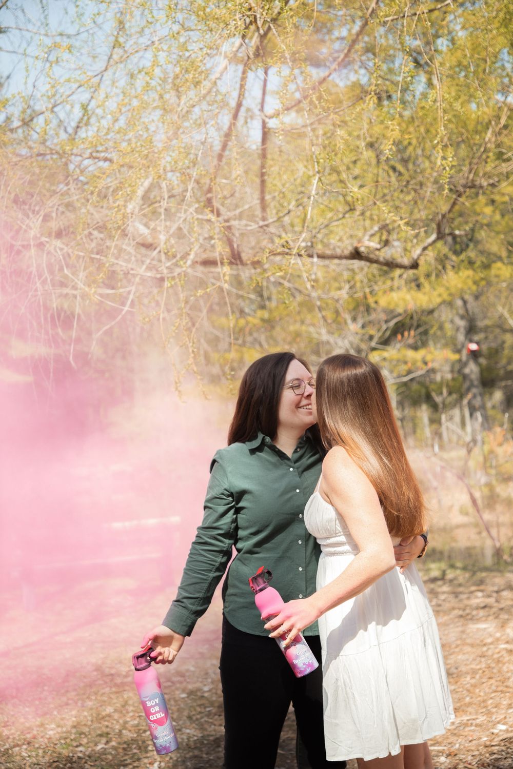 Two women embrace outdoors during a gender reveal, surrounded by pink smoke from handheld cannons among autumn trees.