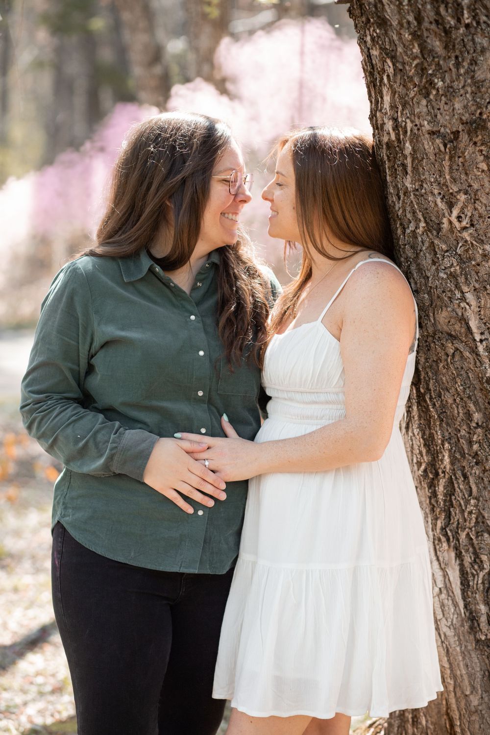 Two women share a romantic moment leaning against a tree with pink blossoms in a sunlit outdoor setting.