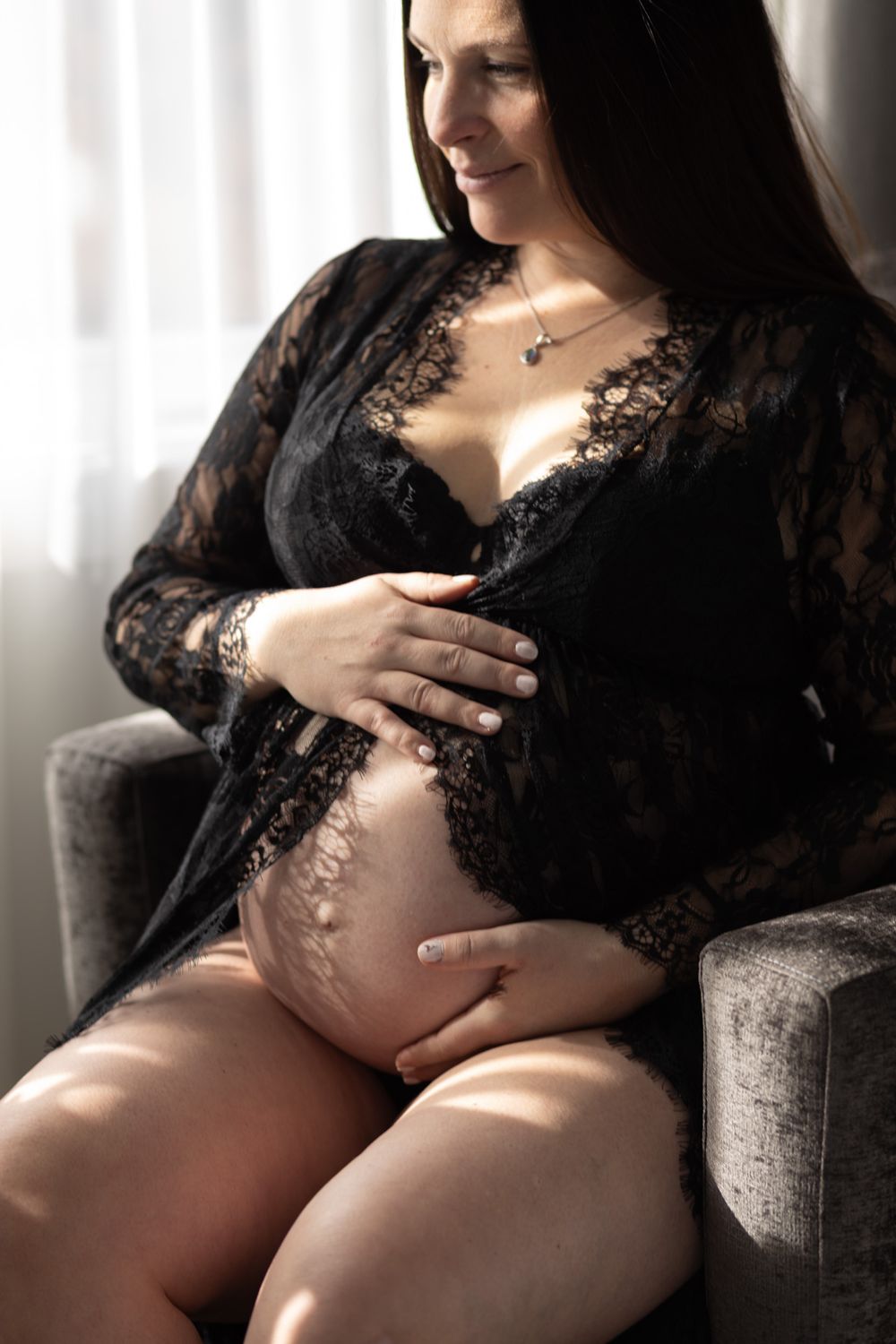 Pregnant woman in black lace robe sitting in chair, cradling baby bump near sunlit window.