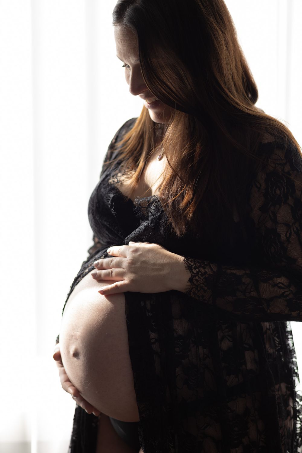 Pregnant woman in black lace robe cradling her baby bump, backlit by window light in an intimate maternity portrait.